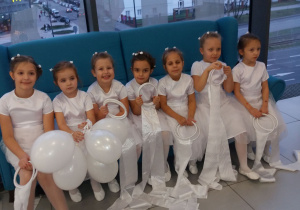 Dziewczynki w białych sukienkach z balonami w rękach i szarfami siedzą na niebieskiej kanapie.